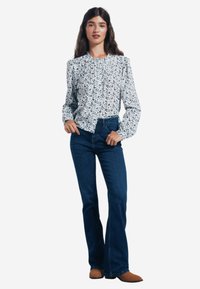 Weißes Blumenbluse mit Rüschenausschnitt und langen Ärmeln, kombiniert mit dunkelblauen Schlagjeans und hellbraunen Knöchelstiefeln. Das Model steht gerade.