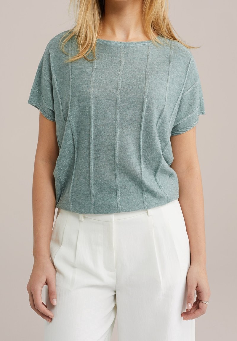 Femme portant un haut en maille manches courtes vert bleu clair et un pantalon plissé blanc taille haute sur un fond uni.