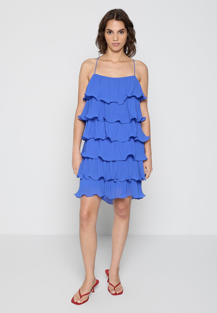 Faltenblaues Kleid mit geschichteten Rüschen, verstellbaren dünnen Trägern und lockerem Schnitt. Getragen mit roten Riemchensandalen, vor einem schlichten Hintergrund.