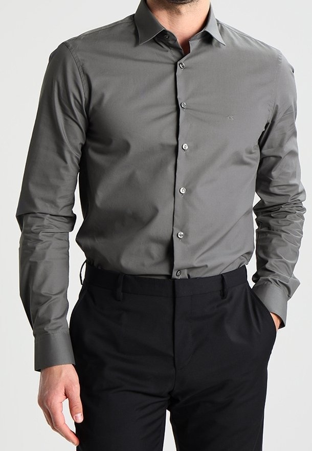 Homme portant une chemise boutonnée gris foncé ajustée rentrée dans un pantalon noir, une main dans la poche et un fond neutre.