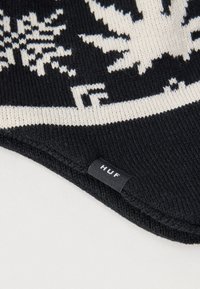 Μαύρο και άσπρο πλεκτό σκουφάκι με σχέδιο νιφάδας χιονιού και μια μικρή μαύρη ετικέτα με την ένδειξη "HUF" στην διπλωμένη άκρη.