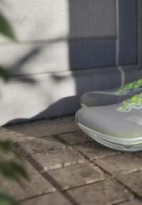 Harmaat lenkkitossut neonvihrein nauhoin, aseteltu kivilaattalattialle varjoisan seinän läheisyydessä.