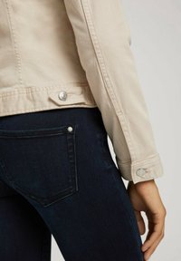 Beige džínsová bunda se stříbrnými prvky, s manžetami na knoflíky, spárovaná s tmavě modrými džíny s detailem zadní kapsy.