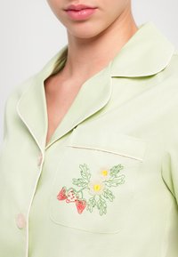 Jasnozielona koszula piżamowa o miękkiej fakturze; posiada kieszonkę z kolorowym haftem w truskawki i kwiaty oraz białą lamówkę wzdłuż krawędzi.