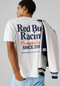 Άνδρας που φοράει λευκό T-shirt με το λογότυπο "Red Bull Racing", κρατάει μπλε ναυτικό και λευκό varsity μπουφάν πάνω στον ώμο του, ανοιχτό γαλάζιο τζιν, γκρι φόντο.
