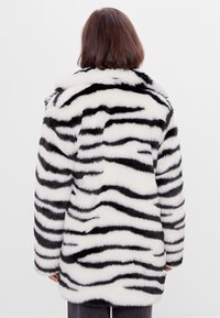 Kabát z umělé kožešiny s černobílým tigřím pruhovaným vzorem, střední délky, širokým límcem a dlouhými rukávy. Měkký, strukturovaný povrch.