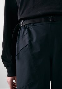 Zwarte broek van gebreide stof, met een elastische tailleband met een gesp, en zijzakken met hoekige naden.