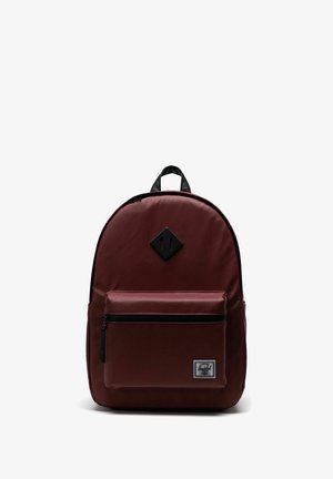 Sac à dos Herschel marron avec poche avant zippée, patch noir en forme de diamant près du haut, et poignée noire contre un fond blanc.