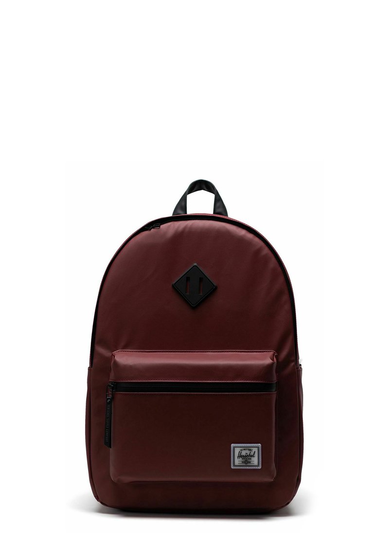 Sac à dos Herschel marron avec poche avant zippée, patch noir en forme de diamant près du haut, et poignée noire contre un fond blanc.