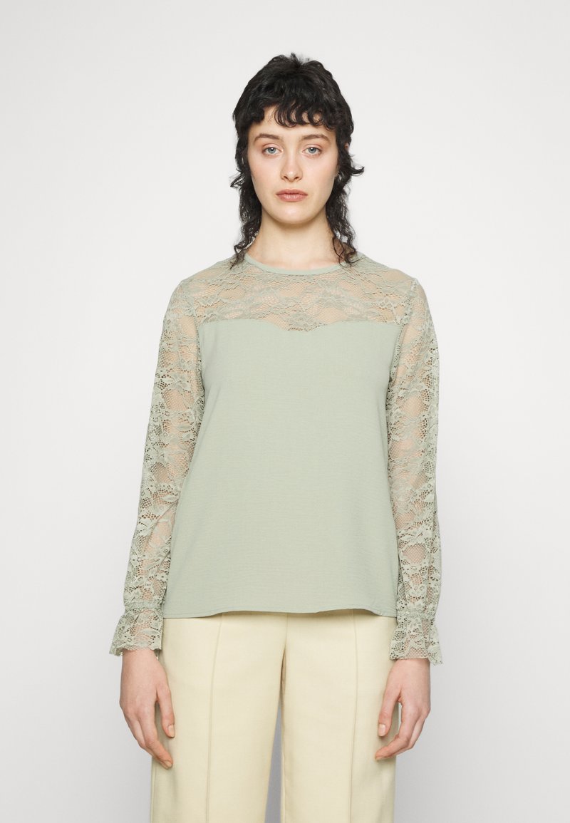 Top verde claro com mangas longas em renda. Textura canelada no corpo principal, padrão floral em renda nas mangas, decote redondo e punhos lânguidos.