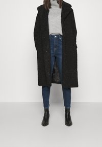 Donna che indossa un lungo cappotto nero a trama sopra un maglione grigio a collo alto, jeans blu a vita alta e stivali neri alla caviglia su uno sfondo semplice.