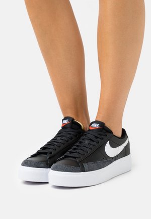 Ben iført sorte Nike-sneakers med hvide swoosh-logoer, sorte snørebånd, grå tåhætte og tykke hvide såler på en ensfarvet baggrund.