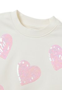 Crème kleurige sweatshirt met meerdere roze iriserende pailletten hartvormen op een zachte stof. Ronde halslijn en geribbelde details.