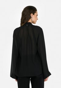 Blouse noire transparente avec des manches longues, un col haut et une fermeture par boutons à l'arrière. Tissu lisse avec une silhouette fluide.