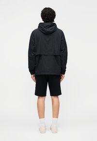 Chaqueta negra con capucha y diseño en capas, fabricada con material ligero. Combinada con pantalones cortos negros y calcetines blancos, usada con zapatos neutros.