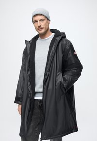 Cămașă de impermeabil neagră, cu căptușeală pufoasă, dotată cu glugă, buzunare frontale și un mic patch cu logo pe mânecă. Caciulă tricotată gri.