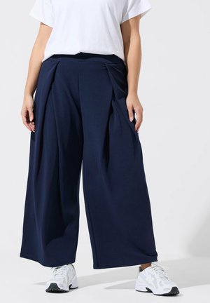 Kobieta w luźnych granatowych spodniach o szerokich nogawkach, białych trampkach i białej koszulce z krótkim rękawem, stojąca na tle gładkiego tła.