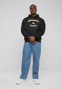 Black hoodie with gold graphic and text, featuring a front pocket and ribbed cuffs, paired with light blue loose jeans and white sneakers.