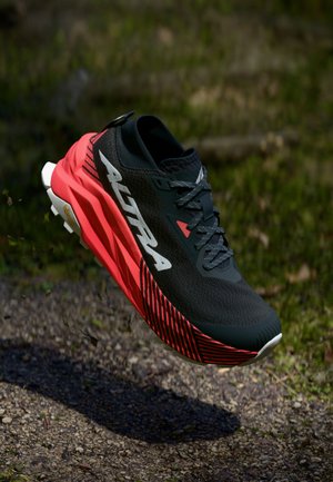 Svart og rød atletisk sko med teksturert overdel, fremtredende rillede såle, reflekterende detaljer, og en Vibram-logo på yttersålen.