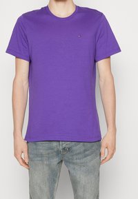 Man som bär en enkel lila t-shirt med korta ärmar och liten logotyp på bröstet, samt blekta blå jeans, mot en ljus bakgrund.