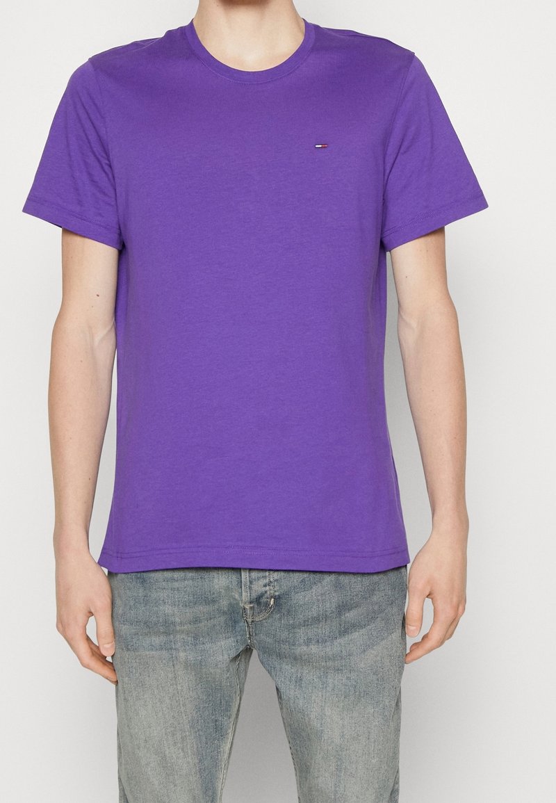 Man som bär en enkel lila t-shirt med korta ärmar och liten logotyp på bröstet, samt blekta blå jeans, mot en ljus bakgrund.