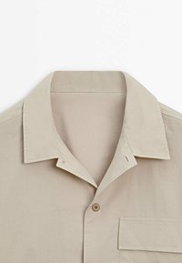 Camicia beige a maniche corte con collo a punta, taschino frontale e un singolo bottone centrale. Tessuto liscio con una sottile texture.