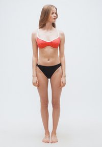 Γυναίκα που στέκεται ξυπόλυτη φορώντας κόκκινο μπικί νι με λευκές τιράντες και μαύρο κάτω μέρος μπικίνι, κοιτάζοντας προς τα αριστερά σε απλό λευκό φόντο.