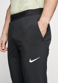 Černé sportovní kalhoty z elastického materiálu s širokým černým pasem označeným "NIKE PRO" a bílým logem Nike swoosh na stehně.