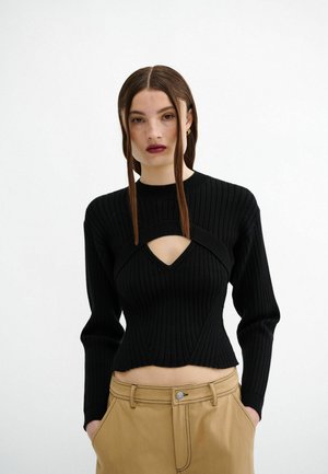 Black ribbed knit sweater with long sleeves and a cut-out detail at the chest, paired with beige high-waisted trousers.