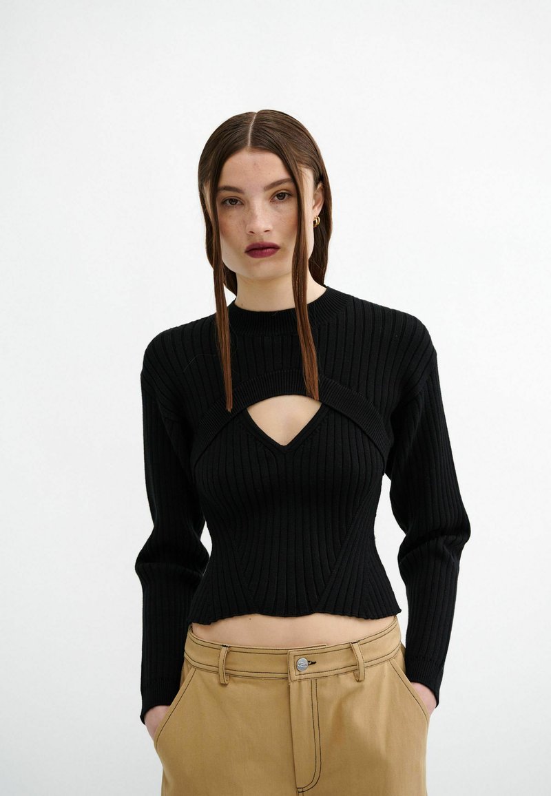 Schwarzer gerippter Strickpullover mit langen Ärmeln und einem Cut-Out-Detail an der Brust, kombiniert mit beigen, hochtaillierten Hosen.