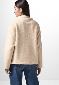 Giacca in pile beige con una texture morbida, vestibilità comoda e colletto. Abbinata a jeans in denim blu a vita alta.