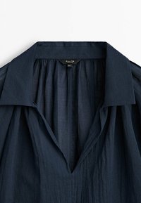 Lekka koszula w kolorze granatowym z szerokim kołnierzem i głębokim dekoltem w kształcie litery V, pokazująca subtelną fakturę i fałdy materiału.