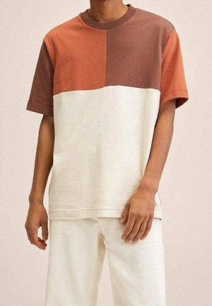 Homme portant un t-shirt à manches courtes color-block avec la moitié supérieure marron et orange et la moitié inférieure crème, associé à un pantalon crème.