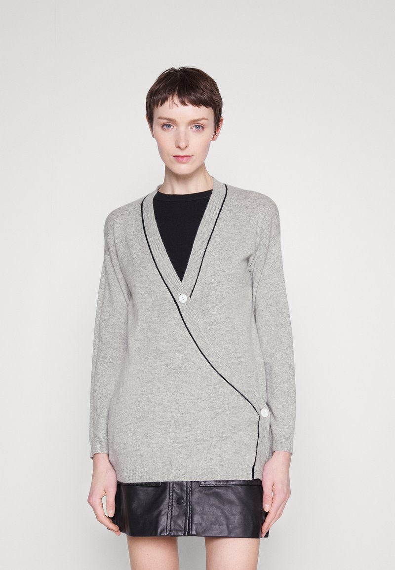 Cardigan gris avec un col en V, bord noir contrastant et fermeture à boutons blancs. Associé à une jupe en cuir noire.