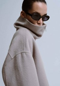 Oversized beige fleece coltrui met een hoge kraag, voorzien van een zijrits, gecombineerd met zwarte ovale zonnebrillen. Soepele textuur.