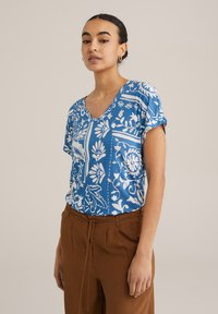 Zils īsām piedurknēm krekls ar baltu ziedu rakstu, V veida kakla izgriezums un brīvs piegriezums, saskaņots ar brūnām biksēm ar aukliņu viduklī.