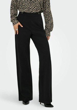 Donna che indossa pantaloni neri a gamba larga, blusa a maniche lunghe con stampa zebrina infilata dentro, tacchi a punta neri e anelli d'argento alle dita.
