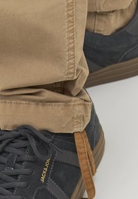 Tmavě šedé tenisky z texturované semišové kůže s gumovými podrážkami, spárované se světle hnědými kalhotami s ohrnutím, které mají viditelné logo značky a tkaničky.