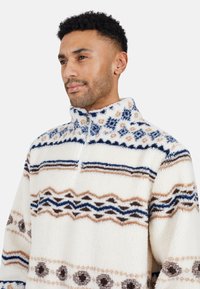 Uomo con capelli ricci corti che indossa un pullover di pile bianco con motivi geometrici blu, marroni e beige e un colletto con zip a metà.