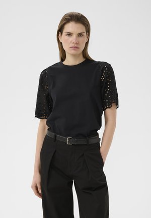 Žena s rovnými hnedými vlasmi má na sebe čiernu blúzku s krátkymi čipkovanými rukávmi a čierne nohavice s opaskom, stojí pred jednoduchým svetlým pozadím.