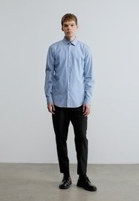 Helehall sinine pika varrukaga särk, millel on ees nupud, kantud mustade pükste ja mustade kingadega. Minimalistlik disain tipuga kraega ja sileda tekstuuriga.