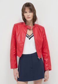 Rote Kunstleder-Bolerojacke mit hohem Kragen, frontalem Reißverschluss und langen Ärmeln, kombiniert mit einem weißen Oberteil und marineblauen Shorts.