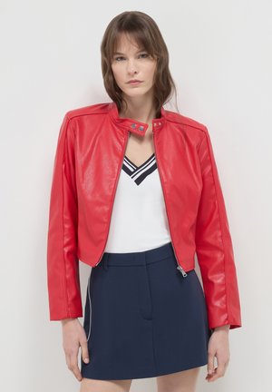 Rote Kunstleder-Bolerojacke mit hohem Kragen, frontalem Reißverschluss und langen Ärmeln, kombiniert mit einem weißen Oberteil und marineblauen Shorts.