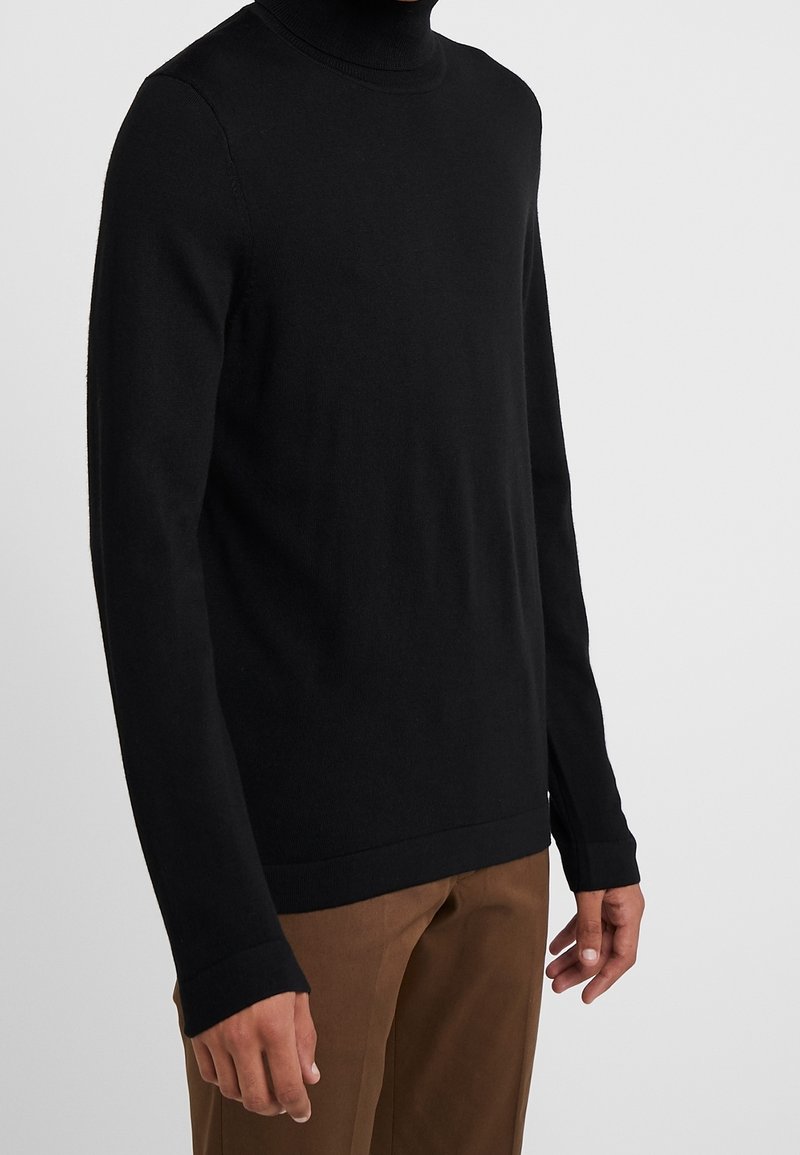 Mann in einem schwarzen Rollkragenpullover mit langen Ärmeln und braunen Hosen vor einem schlichten hellen Hintergrund.