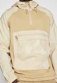 Chaqueta pullover beige con un bolsillo frontal de malla, cierre de media cremallera, capucha y puños elásticos; presenta una marca en la parte inferior derecha.