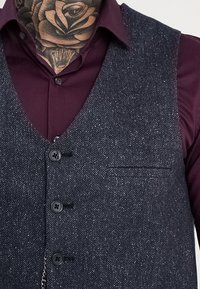 Gilet in tessuto testurizzato a spina di pesce grigio scuro con cinque bottoni neri e una tasca, indossato sopra una camicia bordeaux.