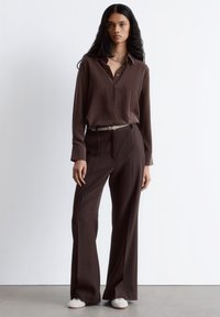 Brown button-up shirt with long sleeves and relaxed fit, paired with matching loose trousers and a light gray belt. Plain white shoes.