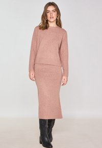 Ensemble en tricot rose pâle comprenant un pull et une jupe assortie avec une texture côtelée, un col rond et une silhouette ajustée, associé à des bottes noires.
