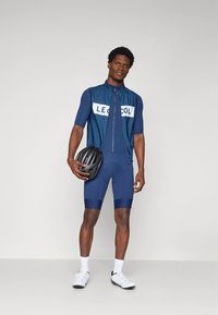 Ναυτικό μπλε ποδηλατικό σύνολο που περιλαμβάνει κοντομάνικο τζέρσεϊ με λευκό λογότυπο, ταιριαστά σορτς με τιράντες και ένα μαύρο κράνος που κρατιέται στο χέρι.