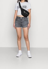 Černá kabelka přes rameno nošená přes bílé pruhované crop top, v kombinaci s šedými džínovými šortkami s roztrhanými okraji a bílými kotníkovými teniskami.